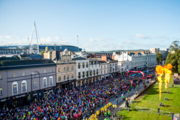 Aeiral view of crowded marathon runners along Cardiff street. Principality Stadium can be seen in the background beyond the street's rooftops. In the foregroud are yellow clothed stewards and flaming pyrotechnics erupting vertically